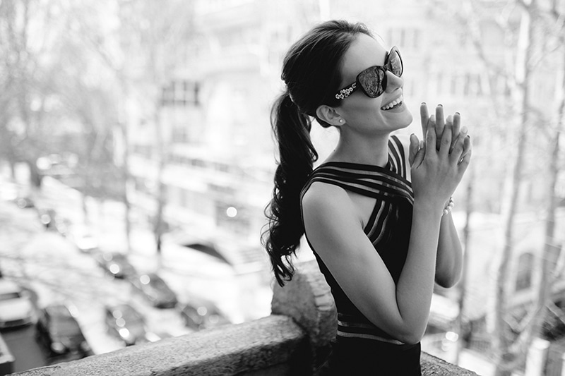 A beautiful,elegant brunette in black sunglasses, sexy black dress, hair ponytail and special features, smiles adorably and looks at one side with hands close to face.Horizontal view on the balcony.